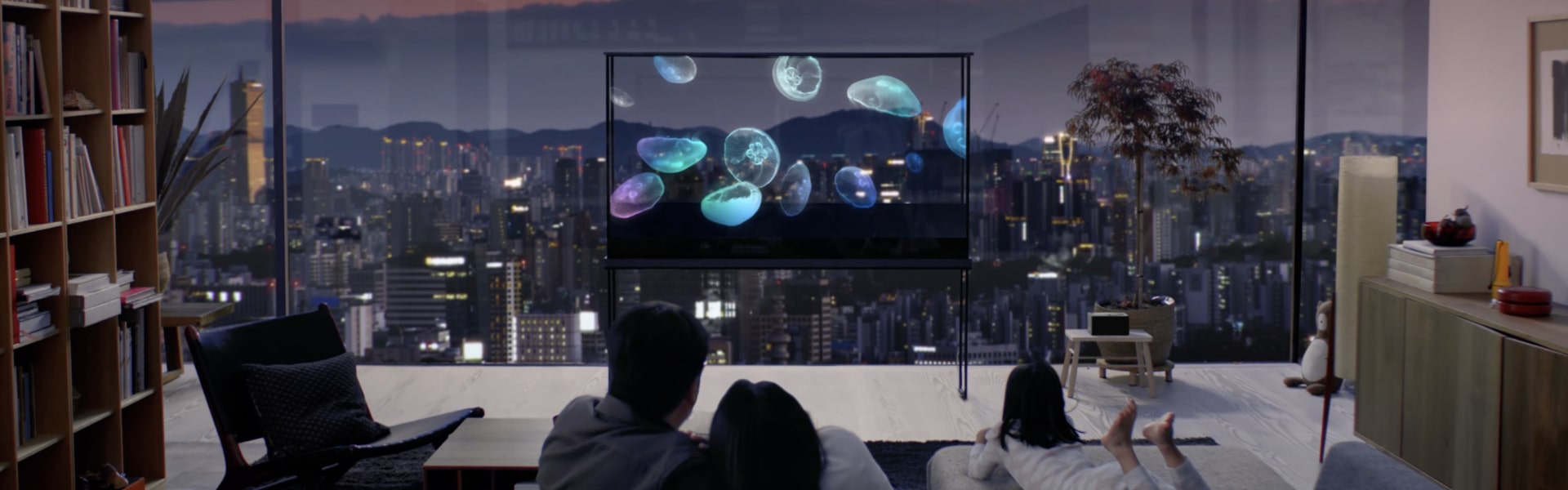 A family enjoys an LG Oled T with a jellyfish floating on it, while the cityscape shines through the transparent screen.