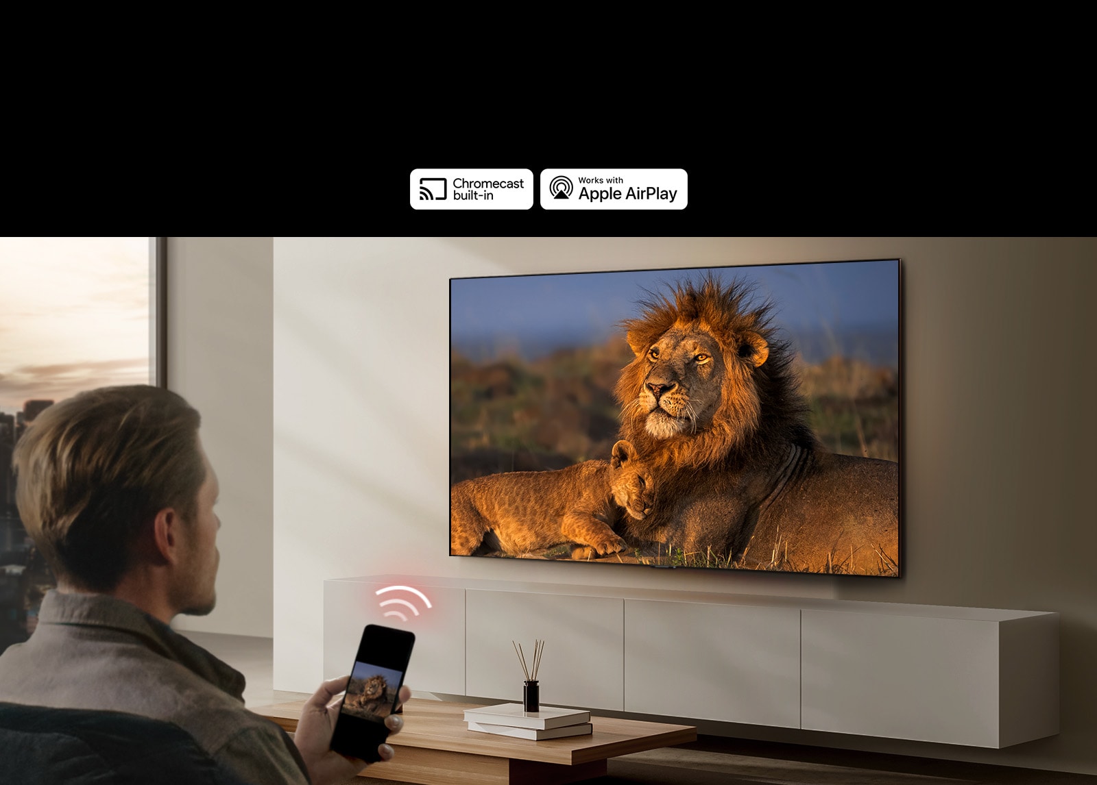 An LG TV mounted on a wall in a living room, displaying a lion and lion cub. A man sits in the foreground with a smartphone in his hand displaying the same image of lions. A graphic of three neon red curved bars is displayed just above the smartphone pointing toward the TV.